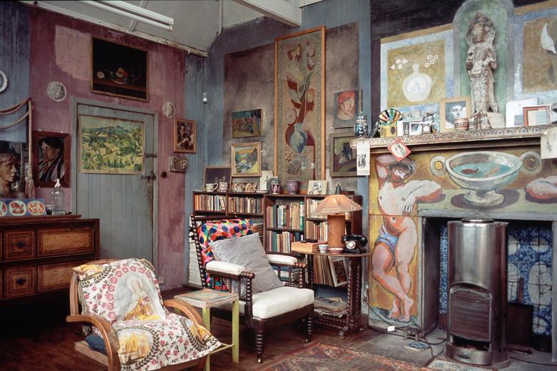 A cluttered corner of an indoor art studio - walls painted in various block colours, two chairs with multicoloured cushions, paintings and sculptures covering the walls. A bookshelf in the background and an unlit fireplace with a tiled and painted facade 