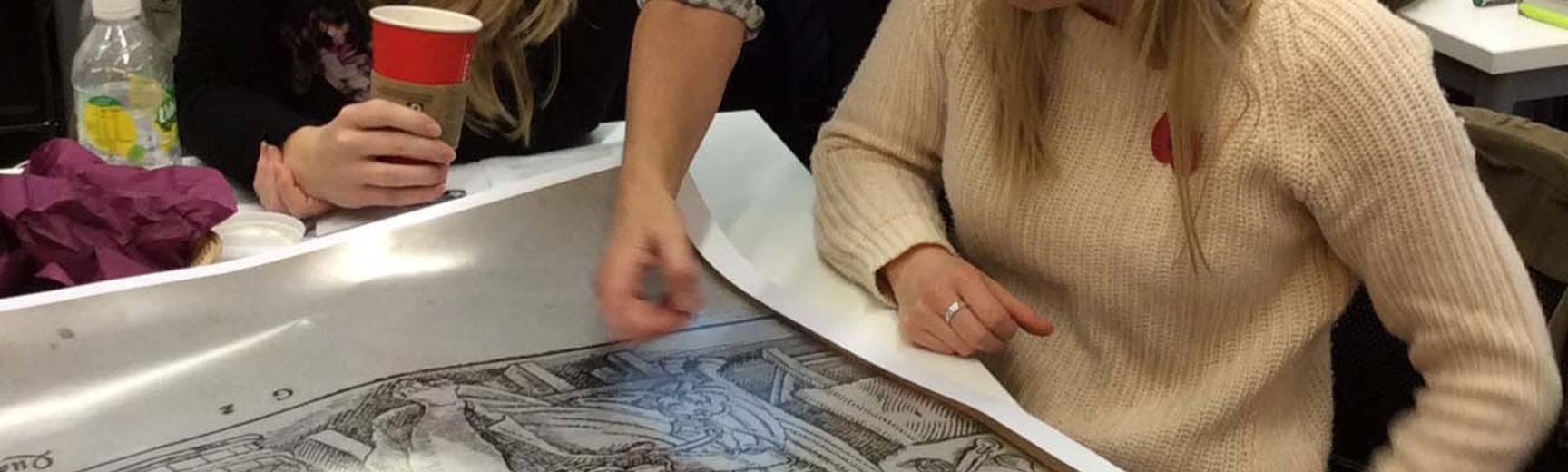 A woman stood between two students points at a poster about midwifery.