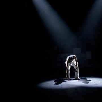 Knowledge Exchange -Theatre A performer under a spotlight touching one hand on the floor of a dark stage