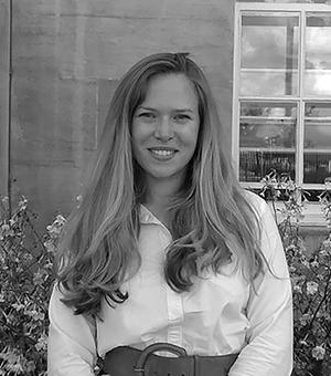 Alice looks at the camera smiling. She has long light brown hair, wears a white shirt and stands in from of the TORCH building. 
